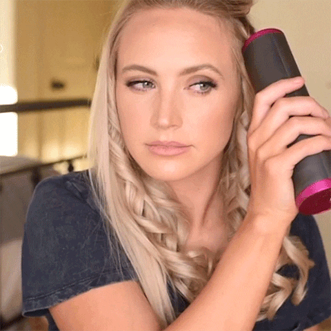 En kvinde med langt blondt hår krøller sit hår med Trådløs Krøllejern - Ledningsfri Automatisk Hair Curler fra Stylemekka.dk, mens hun kigger til siden i et svagt oplyst rum.