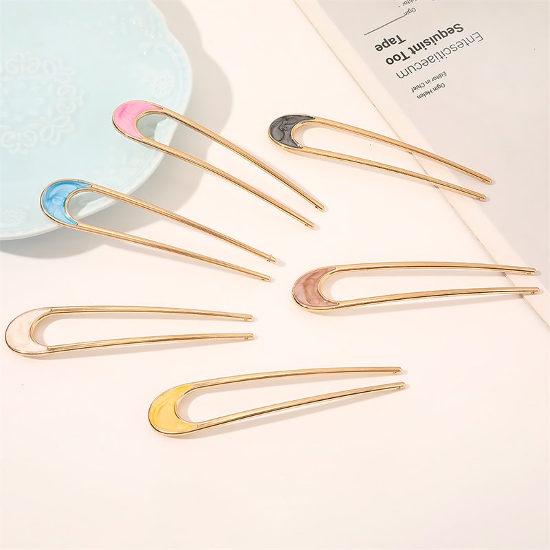 Chris Rubin Alina Fransk Hårnål - Champagne vises med seks guldfarvede hårnåle med emalje i pink, sort, blå, hvid, gul og beige, arrangeret halvcirkelformet på en lys flade ved siden af en blå plade og hvidt papir.