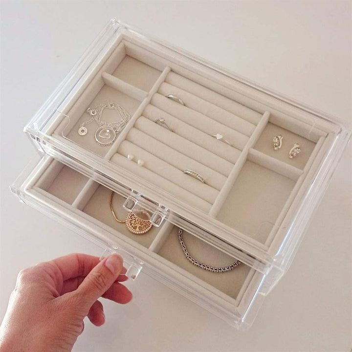 En hånd åbner UNIQ Akryl Smykkeskrin med 3 Skuffer - denne stilfulde UNIQ organizer har et klart akryldesign og beige fløjlsforet interiør, der holder styr på halskæder, armbånd, ringe, øreringe og vedhæng i separate rum.