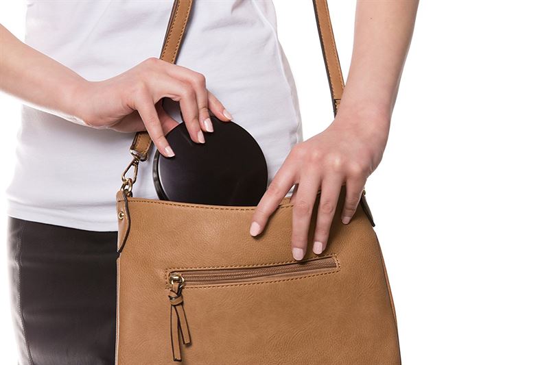 En person i hvid skjorte og sort nederdel lægger en sort pung ned i en brun læderhåndtaske med lynlåslomme ved siden af UNIQ Kompakt Dobbelt Rejsespejl med LED og x10 Forstørrelse (Sort) fra UNIQ på en hvid baggrund.