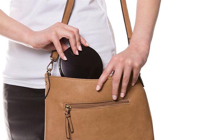 En person i hvid skjorte og sort nederdel lægger en sort pung ned i en brun læderhåndtaske med lynlåslomme ved siden af UNIQ Kompakt Dobbelt Rejsespejl med LED og x10 Forstørrelse (Sort) fra UNIQ på en hvid baggrund.