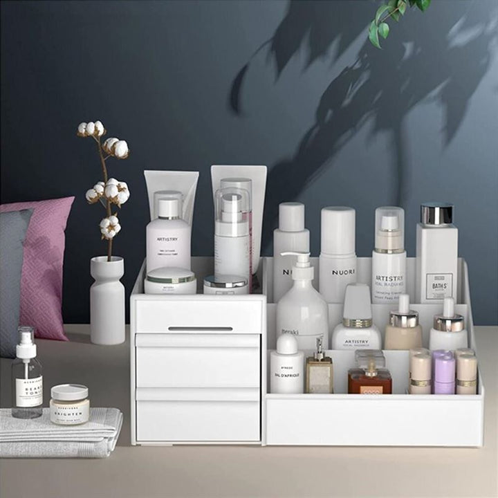 Et ryddeligt toiletbord med UNIQ Kosmetik Organizer | P110 | Hvid by UNIQ og hvide organizere, der viser hudplejeflasker, tuber, krukker og stearinlys med en lille vase med bomuldsstængler på en beige bordplade mod en mørkegrå væg med puder i baggrunden.