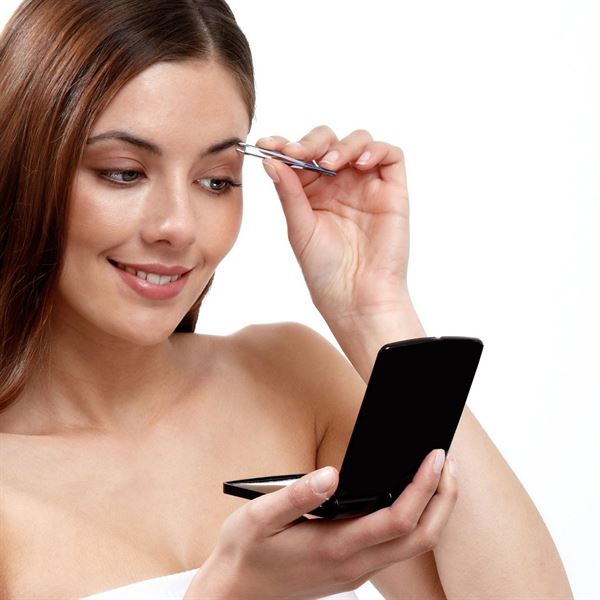 En kvinde smiler, mens hun plukker sine øjenbryn med en pincet og holder UNIQ Lomme- og Makeup Spejl med LED Lys by UNIQ i den anden hånd på en hvid baggrund.