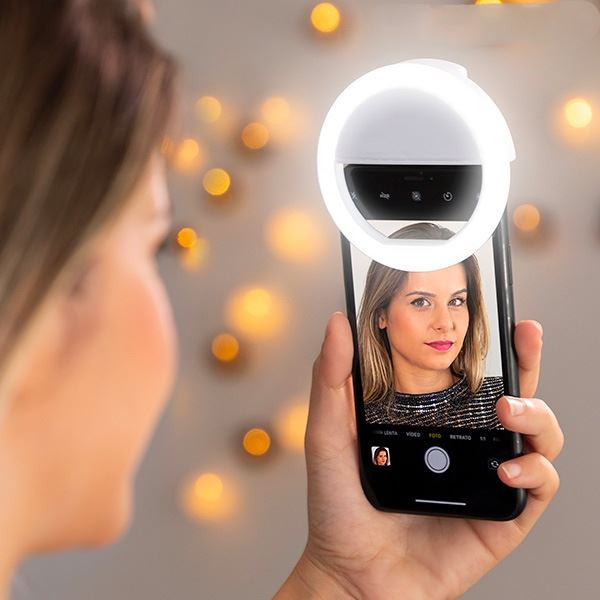 En kvinde tager en glødende selfie med UNIQ Selfie LED Ringlight til smartphones | Genopladelig. Hendes ansigt er oplyst, mens varme, slørede lys i baggrunden skaber en indbydende scene - perfekt til forbedret smartphone-belysning fra UNIQ.