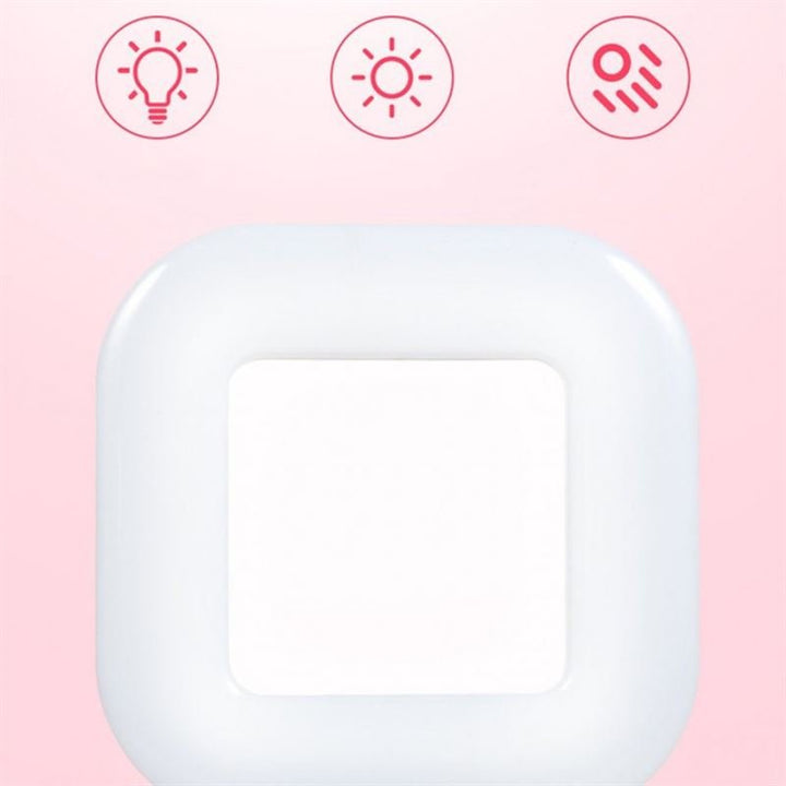 En hvid, firkantet UNIQ Selfie Ringlight til iPhone og Android er centreret på en lyserød baggrund med tre røde ikoner over sig - en pære, en sol og en sensor - som fremhæver dens LED-lysstyrke og bevægelsesdetekteringsfunktioner til smartphones.