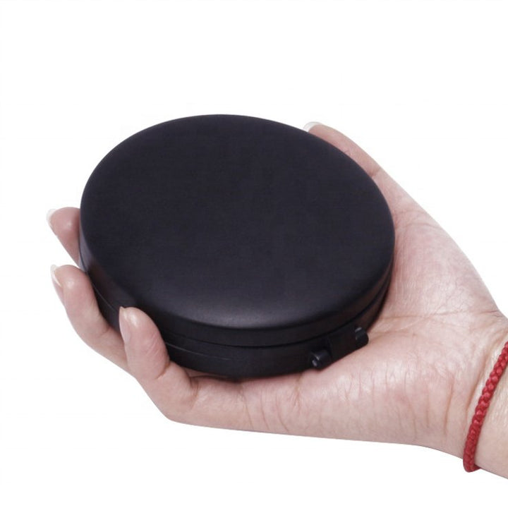 En hånd holder det runde, sorte UNIQ Tri-fold Kompakt Rejsespejl med LED inklusiv forstørrelse. Kompaktspejlet fra UNIQ har en glat overflade, og personen bærer et rødt snorarmbånd om håndleddet.