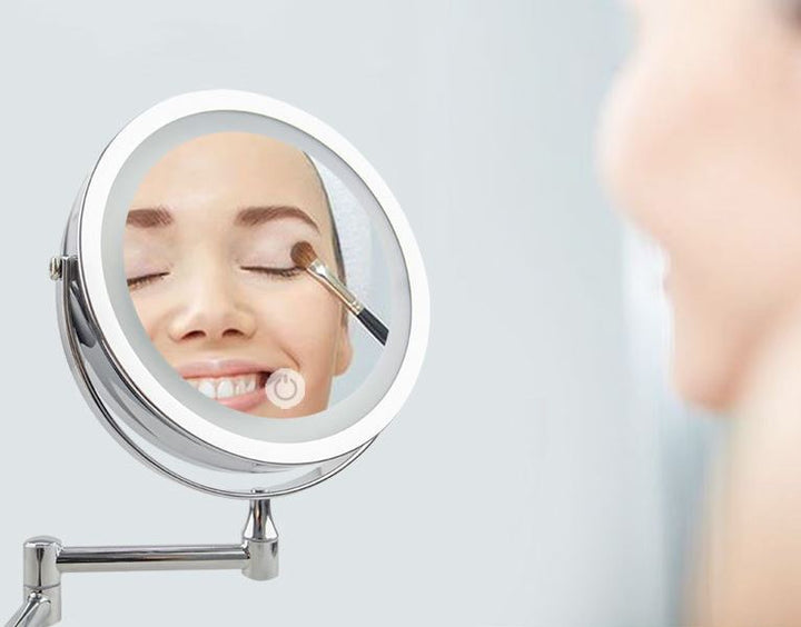 En kvinde smiler, mens hun lægger øjenskygge, reflekteret i UNIQ Vægmonteret LED Kosmetikspejl med leddelt arm og forstørrelse fra UNIQ. Det sølvindrammede makeupspejl giver kraftig LED-belysning, så hendes ansigt er klart synligt.