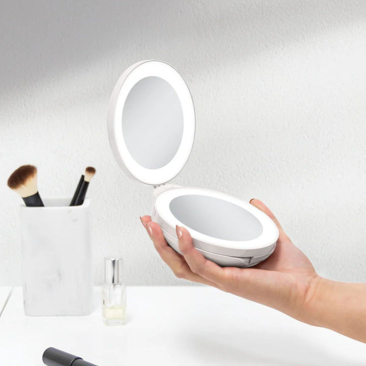 En person holder et åbent UNIQ Kompakt dobbelt rejsespejl med LED (10x forstørrelse) – hvidt. I nærheden ligger nogle makeupbørster i en hvid holder og en lille flaske på en hvid bordplade.