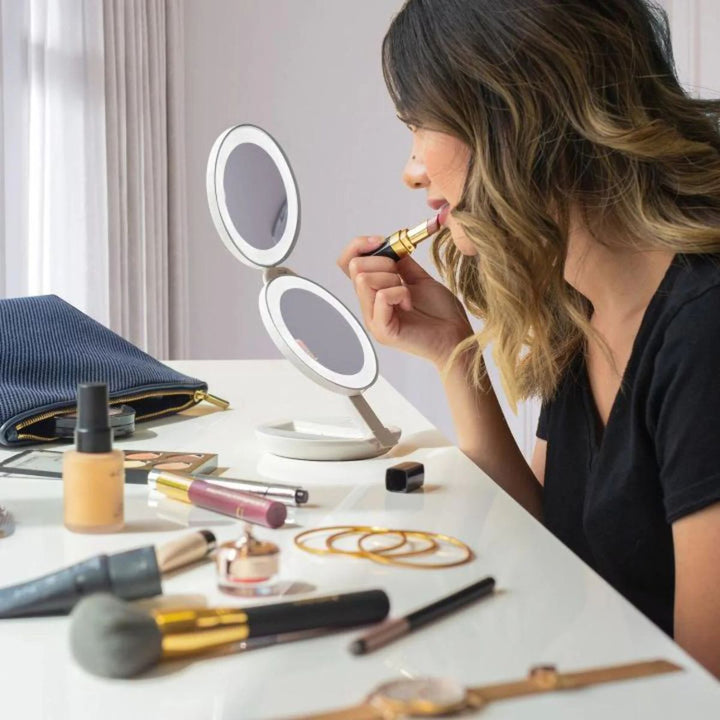En kvinde med skulderlangt hår tager læbestift på, mens hun sidder ved et hvidt bord med et UNIQ Kompakt dobbelt rejsespejl med LED (10x forstørrelse) – Hvid, omgivet af makeup-produkter, børster og smykker.