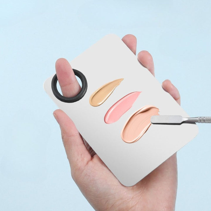 En hånd holder Technique PRO Professional Makeup Mixing Palette, der er fremstillet af rustfrit stål og indeholder cremefarver i beige, lyserød og fersken. En spatel bruges til nem udjævning af foundation på den elegante palet.