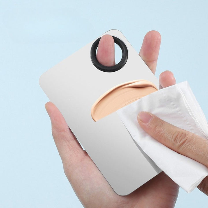 En hånd tørrer Technique PRO Professional Makeup Mixing Palette – der er fremstillet af rustfrit stål og har et praktisk tommelfingerhul – af med et stykke papir. Paletten indeholder en smule beige foundation, hvilket gør den ideel til jævn udtoning af foundation.