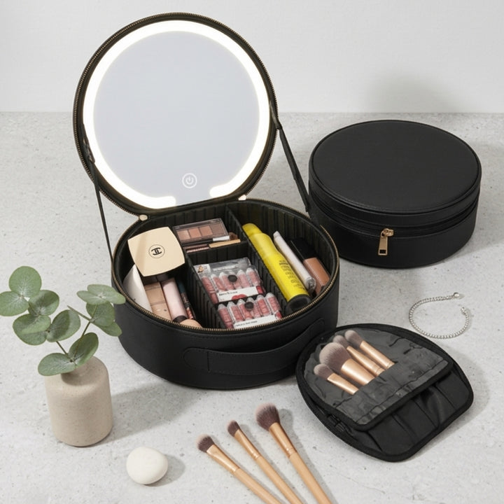 UNIQ Sort LED-makeupspejl med 3-farvet lys og praktisk opbevaring – rejse-makeupkuffert fra UNIQ vises her i åben tilstand, hvor man kan se de pænt organiserede kosmetikprodukter og børster inde i det runde, sorte design med et LED-belyst spejl, der sikrer den perfekte belysning til en fejlfri påføring af makeup. Når rejse-makeupkufferten er lukket, står den på en lys overflade ved siden af makeupbørster, smykker, en plante og en svamp.