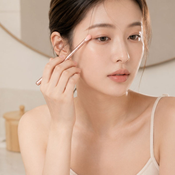 En kvinde lægger øjenskygge på med UNIQ Technique PRO Rose Lens Øjenskyggebørste Sæt, mens hun kigger i et spejl, iført en lys camisole i et blødt oplyst rum.