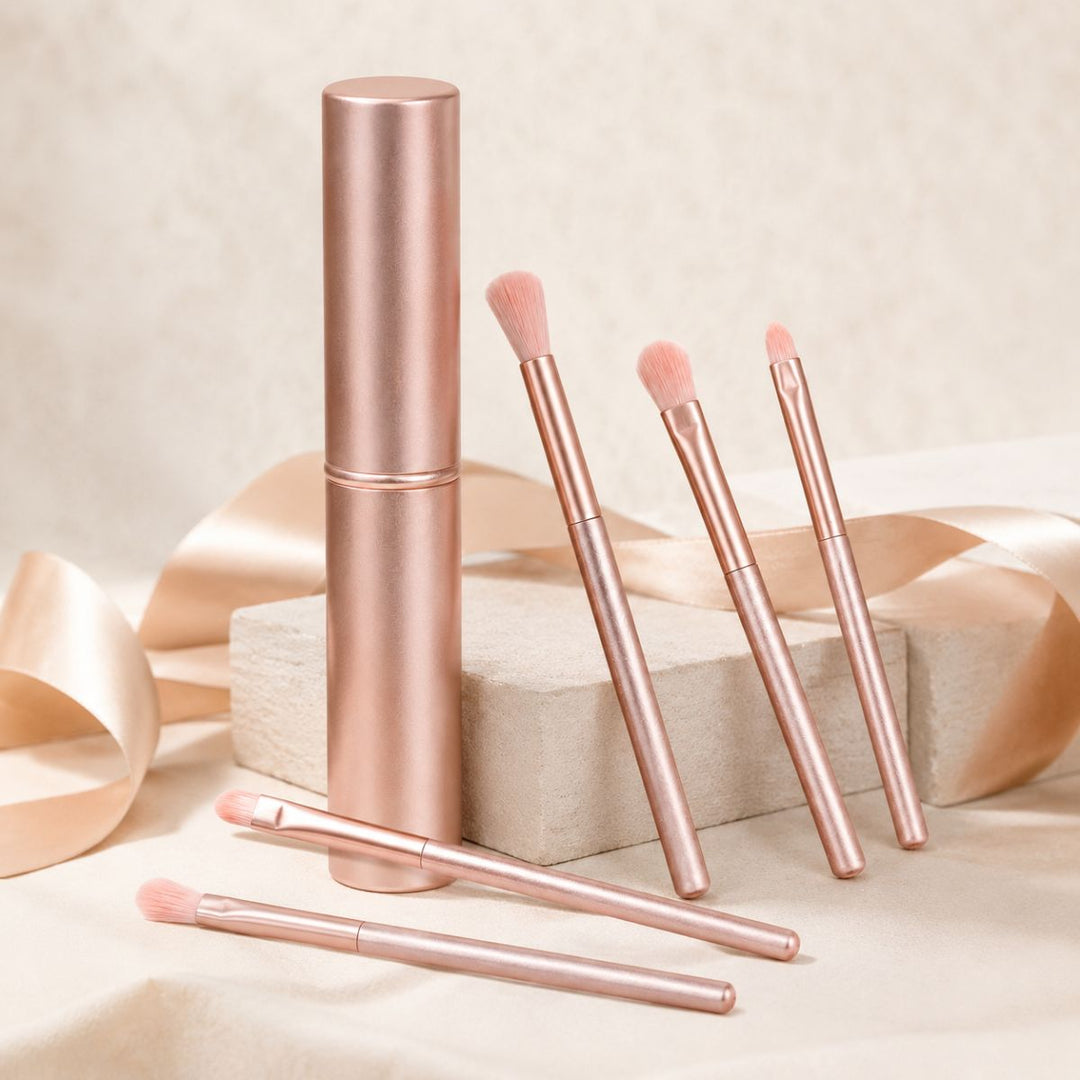 Seks rosaguldbørster fra UNIQ Technique PRO Rose Lens Øjenskyggebørste-sættet og et matchende cylindrisk etui er anbragt på beige blokke med et satinbånd i baggrunden, hvilket skaber et elegant og blødt udtryk.