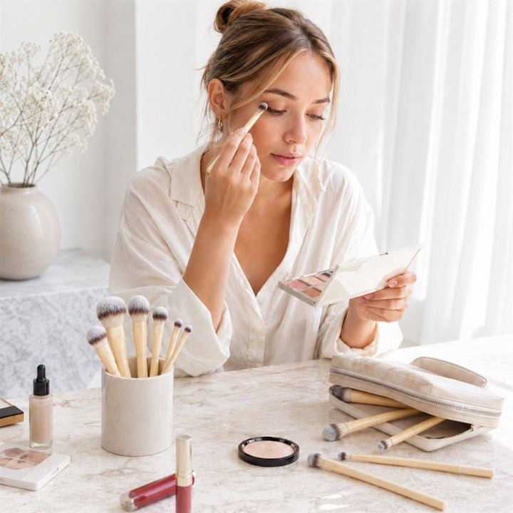 En kvinde i en hvid bluse sidder ved et bord og lægger øjenskygge på med en pensel, mens hun bruger et håndspejl. På bordet ligger der forskellige makeup-produkter samt »Technique PRO Buttercream Makeupbørste Sæt – 10 makeupbørster« fra Technique PRO.