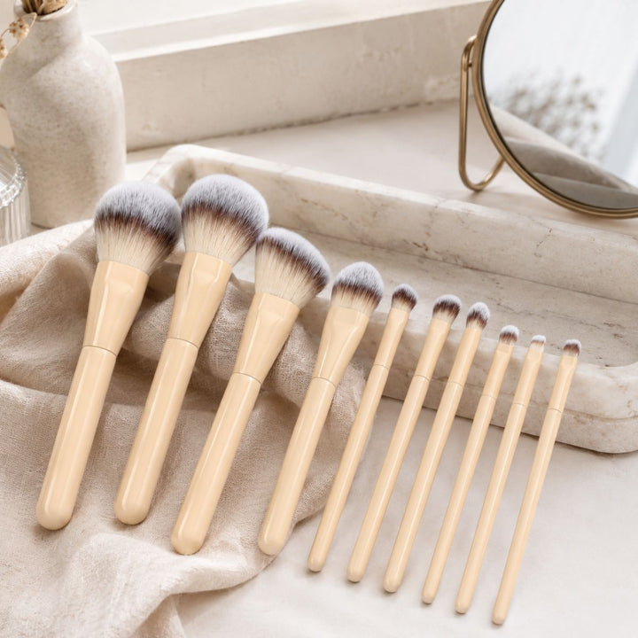 Et sæt bestående af ti Technique PRO Buttercream Makeupbørste Sæt makeupbørster med beige skafter i forskellige størrelser er pænt anbragt på et marmorfad ved siden af et spejl og dekorative vaser på en lys overflade.