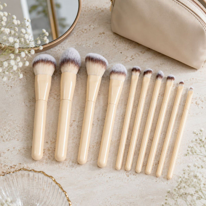 Et sæt bestående af ti Technique PRO Buttercream-makeupbørster, hver med elegante beige skafter og i forskellige størrelser, er pænt anbragt på en beige overflade ved siden af en kosmetikpung, et spejl og dekorative blomster – hvilket gør dette Technique PRO Buttercream-makeupbørstesæt til det perfekte valg for alle, der søger et stilfuldt og funktionelt makeupbørstesæt.