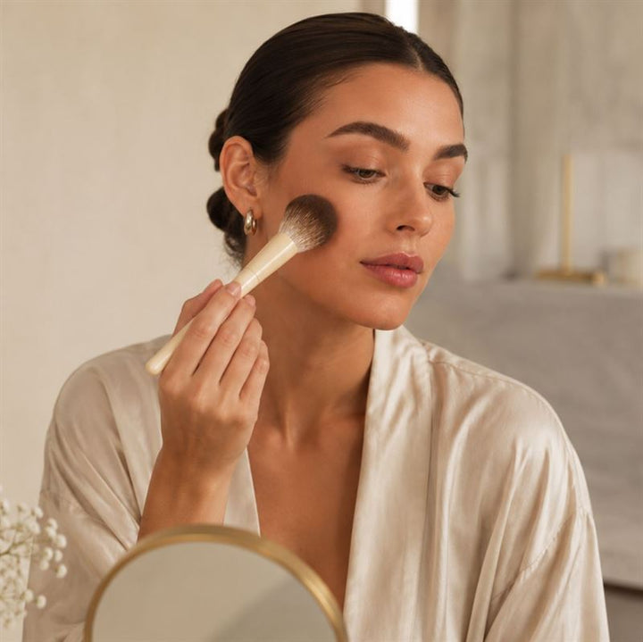 En kvinde i en silke-morgenkåbe lægger makeup på kinden med en pensel fra Technique PRO Vanilla Makeupbørste Sæt – 14 makeupbørster, mens hun kigger i et rundt spejl i et blødt oplyst rum.