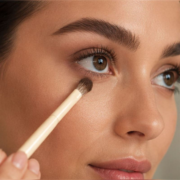 En person lægger makeup under øjnene med en lille børste fra Technique PRO Vanilla Makeupbørste Sæt – 14 makeupbørster fra Technique PRO. Billedet fokuserer på øjenområdet og fremhæver glat hud, velplejede øjenbryn og et naturligt makeup-look.