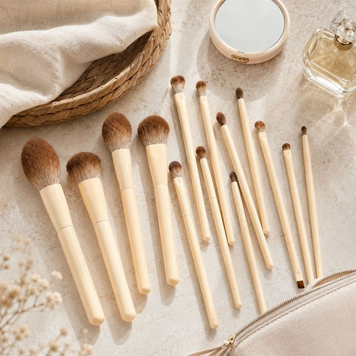 Et sæt på fjorten Technique PRO Vanilla-makeupbørster med beige skafter, pænt anbragt på en lys overflade ved siden af et lille rundt spejl, en parfumeflaske, en flettet måtte og en beige makeup-taske – perfekt til at løfte din makeuprutine.