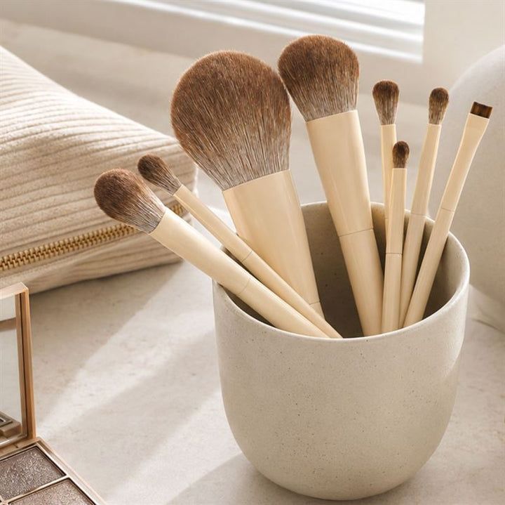 Et sæt bestående af fjorten makeupbørster med beige håndtag fra Technique PRO, elegant anbragt i en lys keramisk skål på en beige overflade, hvor sollyset strømmer ind, ledsaget af en matchende beige taske med lynlås i baggrunden. Technique PRO Vanilla Makeupbørste-sættet er perfekt til nemt at skabe fejlfri looks.