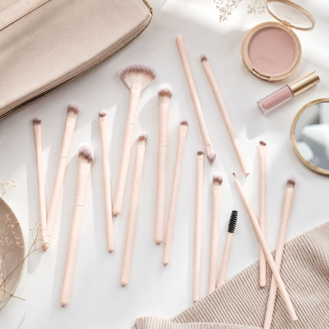 Et sæt Technique PRO Keen Eyeshadow, Eyebrow & Detailing Makeupbørste Sæt – 16 makeupbørster fra Technique PRO er anbragt på en hvid overflade med rouge, en makeup-taske, lipgloss, et spejl og en sweater i nærheden – perfekt til at skabe fejlfri øjenmakeup-looks.