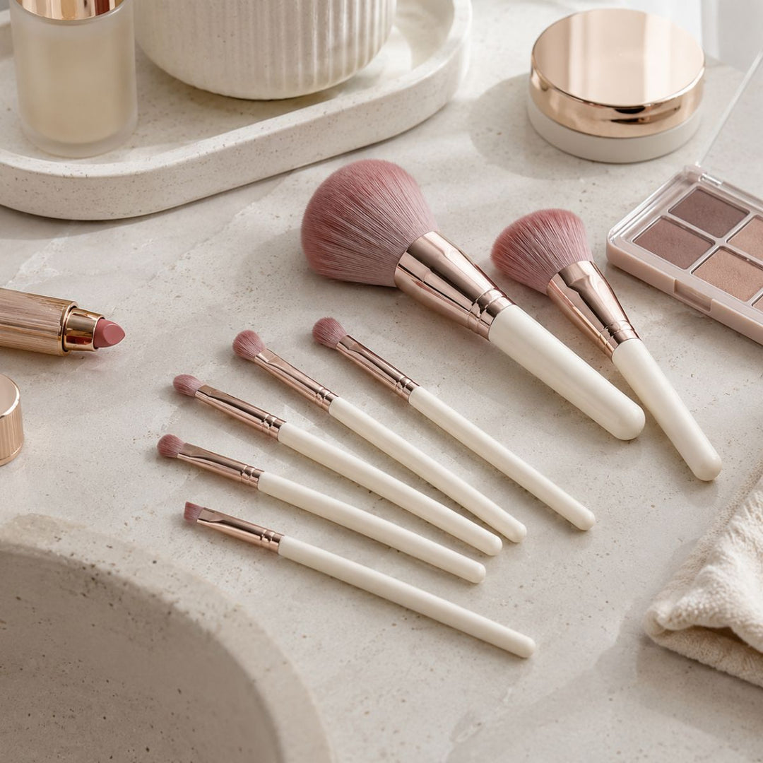 Et sæt bestående af syv Technique PRO Dusty Magenta-makeupbørster er anbragt på en beige bordplade med hvide skafter og rosaguld-ringe, omgivet af makeup-produkter, herunder en læbestift, en øjenskyggepalet og en pudderdåse.