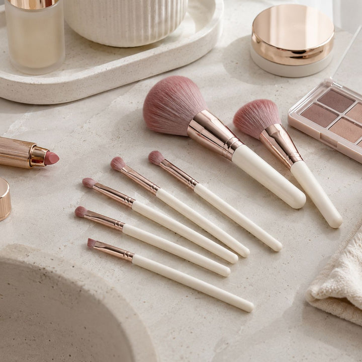 Et sæt bestående af syv Technique PRO Dusty Magenta-makeupbørster er anbragt på en beige bordplade med hvide skafter og rosaguld-ringe, omgivet af makeup-produkter, herunder en læbestift, en øjenskyggepalet og en pudderdåse.
