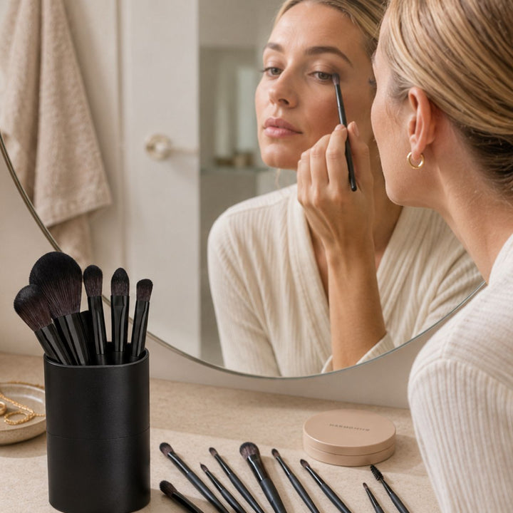 En kvinde lægger øjenskygge på med en pensel fra sit »Technique PRO Raven Makeupbørste Sæt – 18 makeupbørster«, mens hun kigger i et rundt badeværelsesspejl. Technique PRO-makeupbørster, en beholder og pudder står på bordpladen foran hende. Hun har en cremefarvet trøje på og en guldringørering.