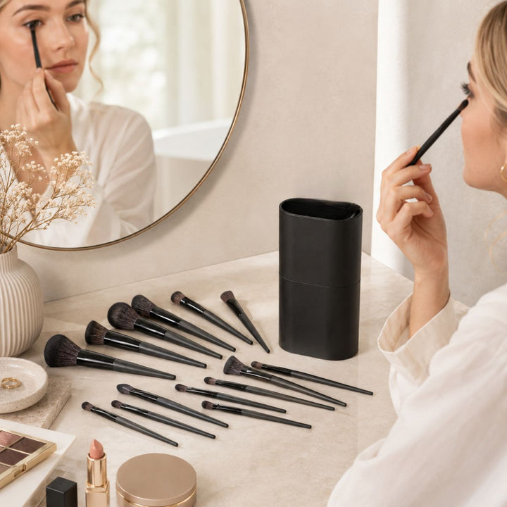 En person lægger makeup med en pensel, mens vedkommende kigger i et rundt spejl. Forskellige makeup-pensler og kosmetik, herunder det elegante »Technique PRO Raven Makeupbørste Sæt – 18 makeupbørster« fra Technique PRO, er pænt opstillet på toiletbordet ved siden af en sort penselholder.