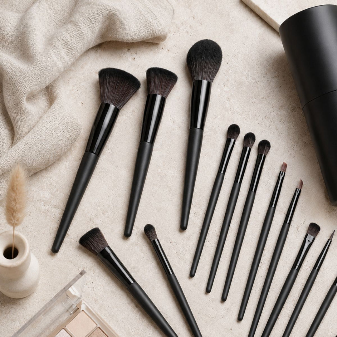 Et Technique PRO Raven-makeupbørstesæt – bestående af 18 makeupbørster med elegante sorte skafter – er pænt anbragt på en beige overflade, ledsaget af en klud, en lille vase og en makeup-palet, der står i nærheden.