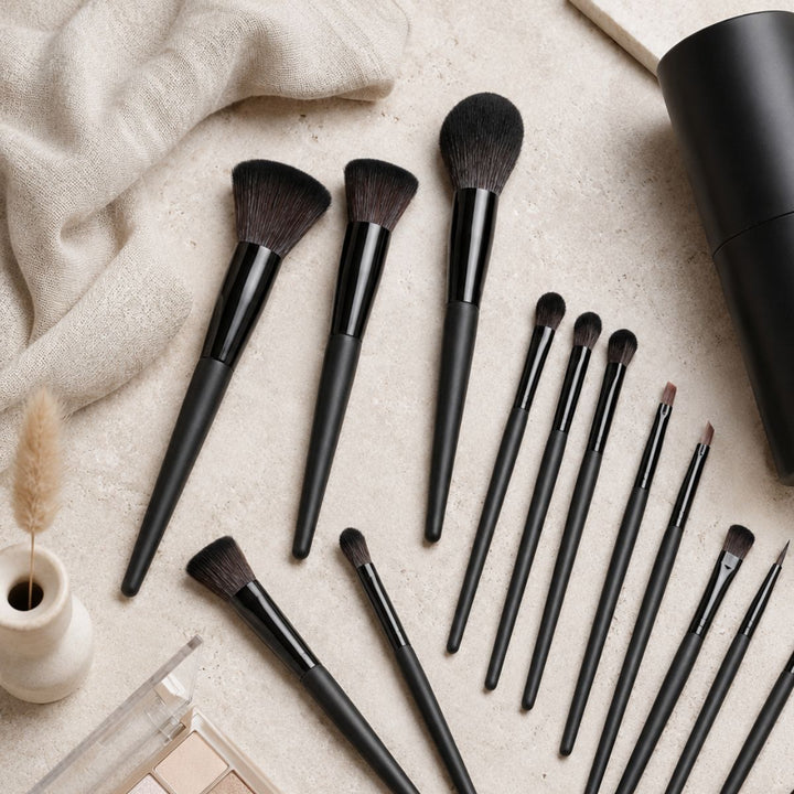 Et Technique PRO Raven-makeupbørstesæt – bestående af 18 makeupbørster med elegante sorte skafter – er pænt anbragt på en beige overflade, ledsaget af en klud, en lille vase og en makeup-palet, der står i nærheden.