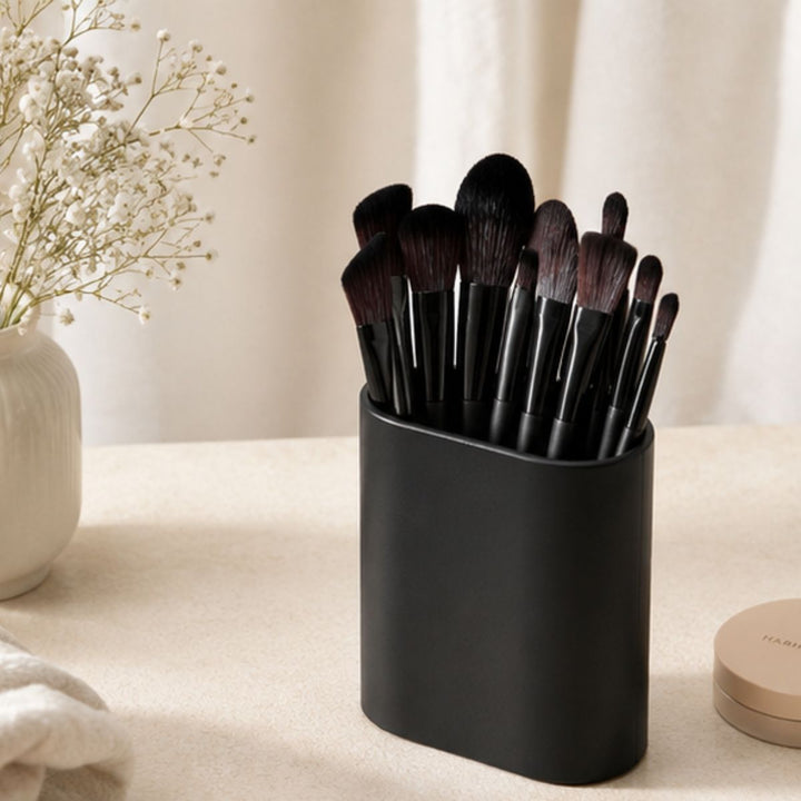 Technique PRO Raven Makeupbørste Sæt – 18 makeupbørster fra Technique PRO, med sorte skafter og præsenteret i en elegant sort holder, står på en beige overflade ved siden af en vase med gipskrydderblomster og en rund, beige pudderdåse. I baggrunden hænger cremefarvede gardiner.