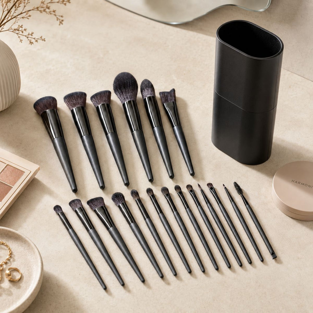 Technique PRO Raven-makeupbørstesættet, der består af 18 professionelle makeupbørster med elegante sorte skafter, er pænt anbragt på en beige overflade ved siden af en sort børsteholder, en makeup-palet, en pudderdåse, smykker og en hvid vase.