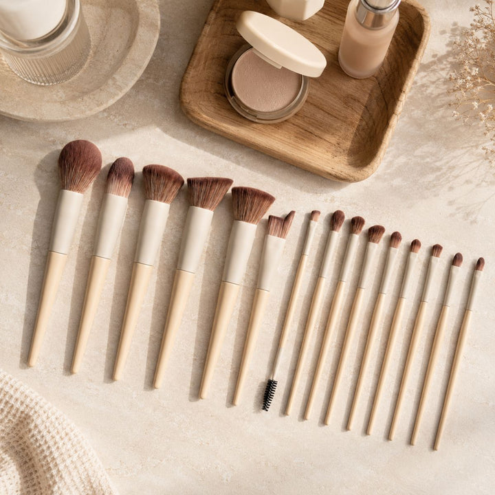 Et 18-delt makeupbørstesæt, Technique PRO Dover Makeupbørste Sæt fra Technique PRO, med beige håndtag, ligger pænt opstillet på en lys overflade ved siden af ansigtspudder, foundation og andre ansigtsmakeupprodukter.