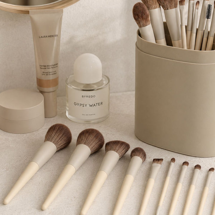 Et sæt Technique PRO Dover Makeupbørste Sæt – bestående af 18 makeupbørster med beige skafter, en Laura Mercier-tube og en Byredo Gypsy Water-parfumeflaske – er pænt anbragt på en lys overflade ved siden af en matchende beige Technique PRO-holder til makeupbørstesættet.