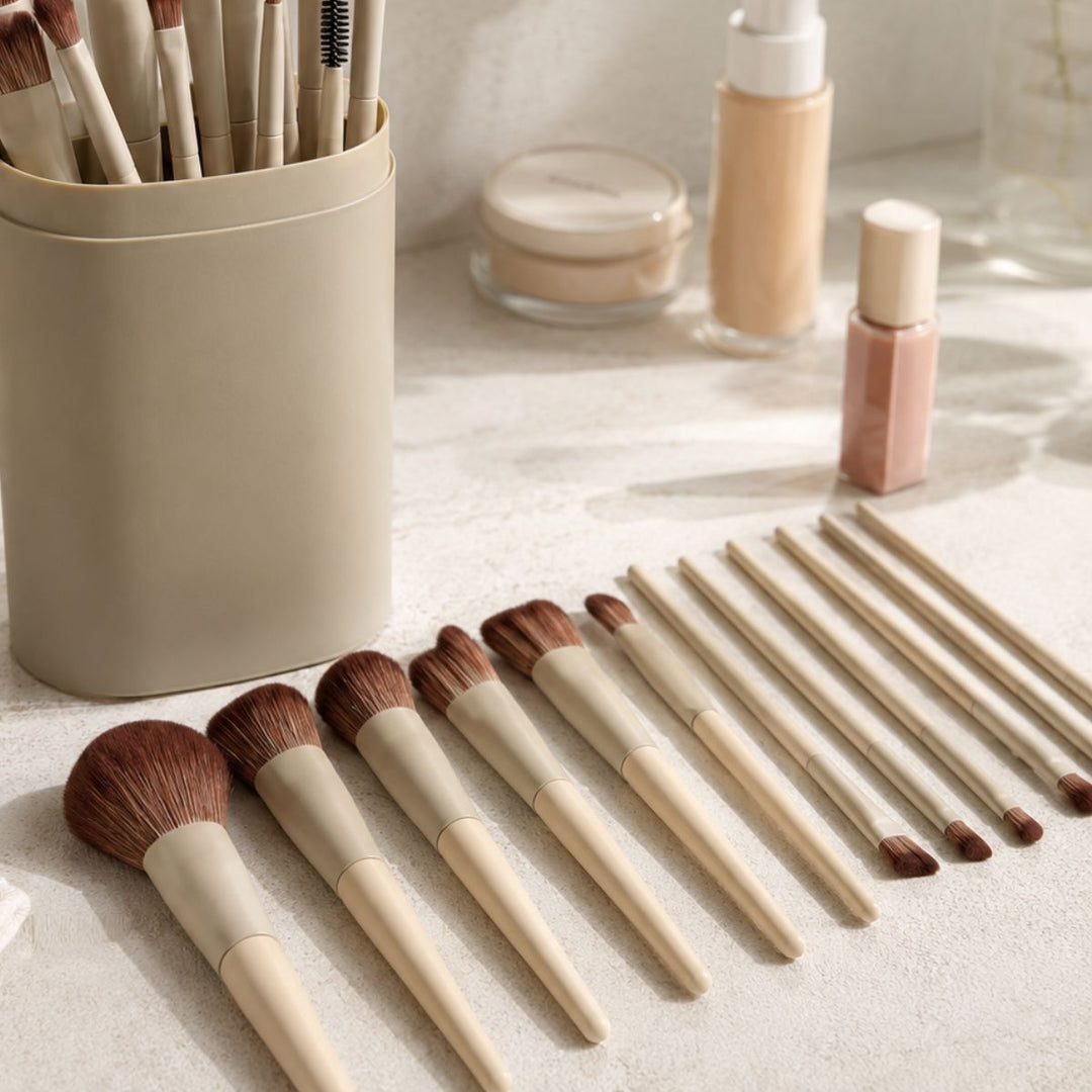 Et sæt Technique PRO Dover Makeupbørste Sæt – 18 makeupbørster i forskellige størrelser – er anbragt på en beige overflade, mens der i baggrunden står en beige beholder med yderligere Technique PRO-børster og flasker med ansigtsmakeupprodukter.