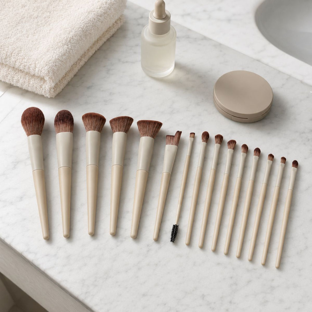 Atten Technique PRO-makeupbørster fra Technique PRO Dover Makeupbørste-sættet, med beige håndtag, en spoolie-børste, en dråbeflaske, et kompakt etui og et sammenfoldet håndklæde, er anbragt på en hvid marmor-badeværelsesbordplade tæt på en håndvask – et elegant sæt, der er perfekt til at skabe fejlfri ansigtsmakeup.