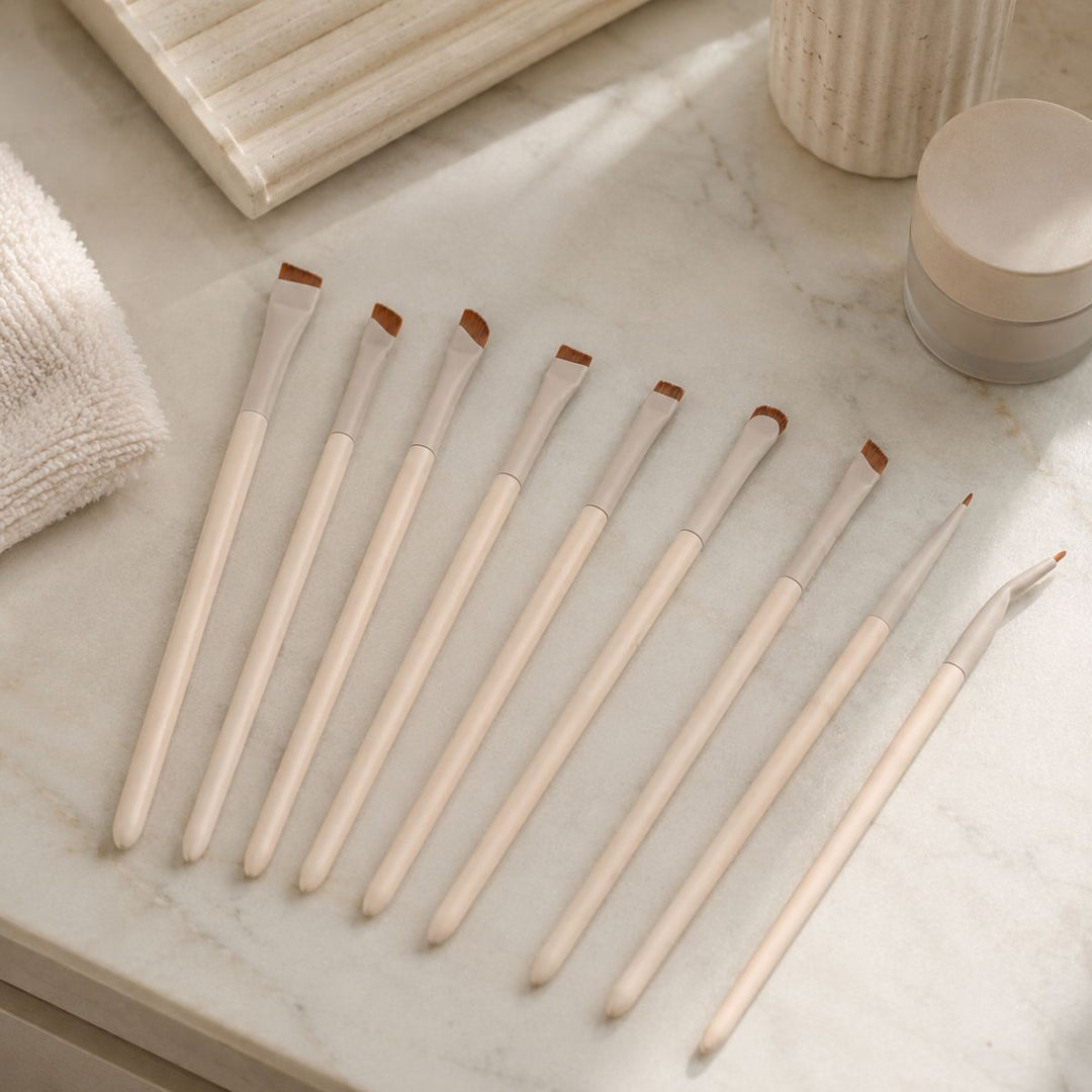 Nine Technique PRO Flick Eyeliner, Bryn & Detail Makeupbørste Sæt – 9 makeupbørster ligger pænt opstillet på en marmorbordplade, hvor deres beige skafter og vinklede børstehår er omgivet af håndklæder og kosmetikbeholdere, herunder et udvalg af præcisions-eyelinerbørster fra Technique PRO.