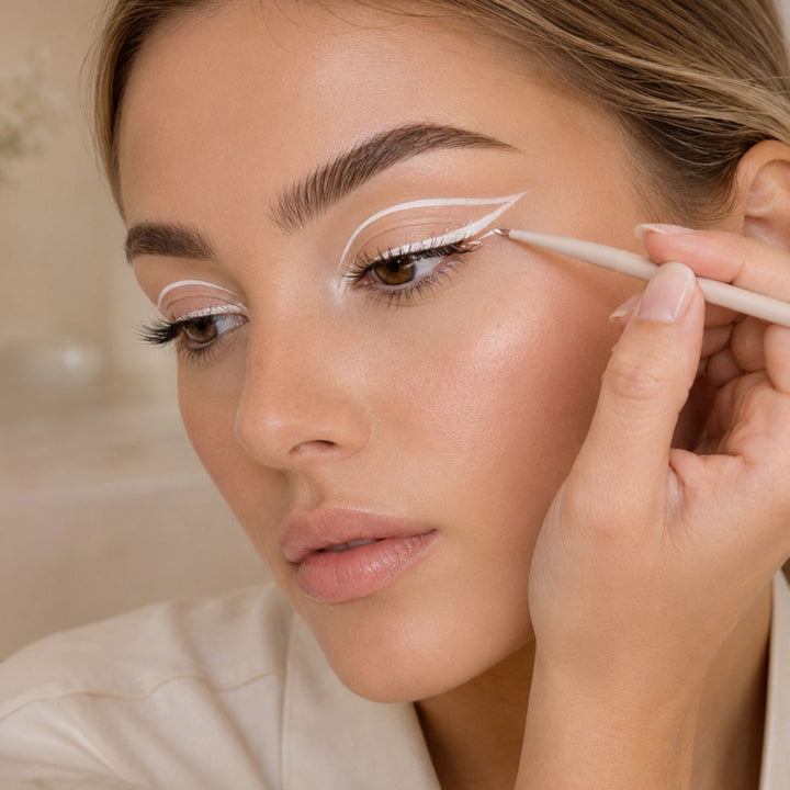 En person bruger Technique PRO Flick Eyeliner, Bryn & Detail Makeupbørste Sæt til at påføre hvid, grafisk eyeliner på det øverste øjenlåg og opnår dermed et markant og kunstnerisk look med dobbelt vinge. Nærbilledet fremhæver præcisionen i påføringen af makeup og de perfekt plejede øjenbryn og viser, hvordan professionelle redskaber som dem fra Technique PROs sæt med 9 makeupbørster – især brynbørsten – er med til at skabe fejlfri resultater.