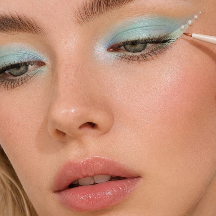Nærbillede af en person med blå øjenskygge, eyeliner med vingeformet spids og små perleagtige accenter, der påføres det ydre øjenområde ved hjælp af en af de præcise makeupbørster fra Technique PRO Flick Eyeliner, Bryn & Detail Makeupbørste Sæt fra Technique PRO. Huden fremstår fugtig, og læberne er glansfulde.