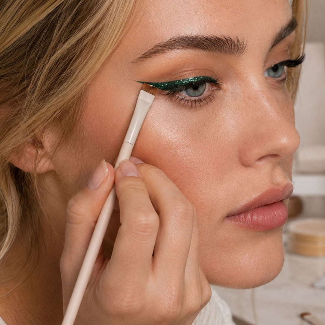 En kvinde bruger Technique PRO Flick Eyeliner sammen med en tynd, vinklet pensel fra Technique PRO Bryn & Detail Makeupbørste Sæt til at påføre grøn, glitrende eyeliner, hvorved hun omhyggeligt skaber en skarp, udstrakt effekt på sit øverste øjenlåg. Hendes hud fremstår glat, og hendes læber har en naturlig, glansfuld finish.