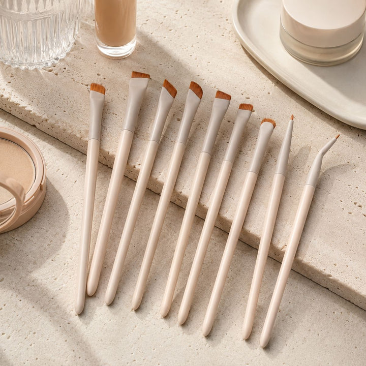 Ni makeupbørster med beige skafter og vinklede børstehår fra Technique PRO Flick Eyeliner, Bryn & Detail Makeupbørste-sættet er pænt anbragt på en struktureret beige overflade, omgivet af kosmetik såsom foundation, pudder og krukker på bakker. Dette elegante sæt fra Technique PRO indeholder uundværlige redskaber som eyelinerbørster og brynbørster til at skabe fejlfri beauty-looks.