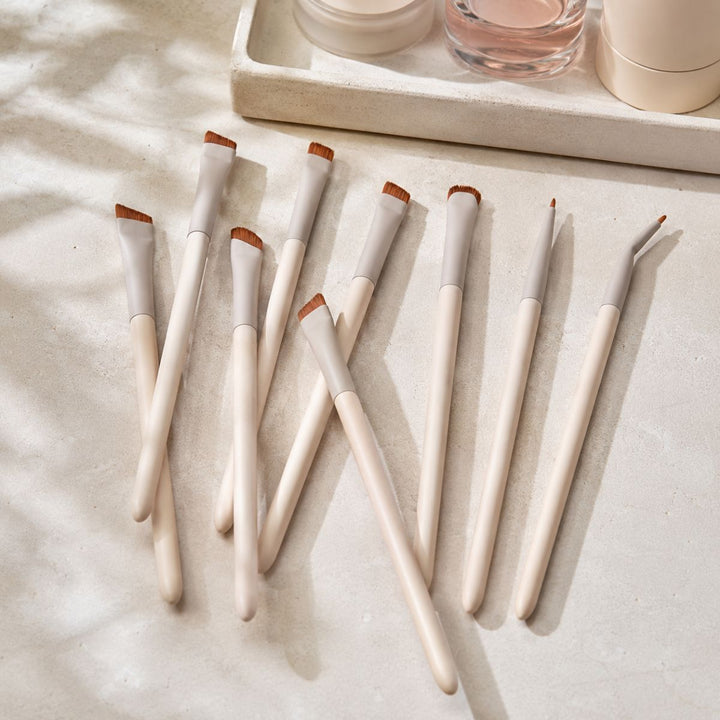 Ni makeupbørster fra Technique PRO Flick Eyeliner, Bryn & Detail Makeupbørste Sæt, med hvide skafter og brune børstehår – herunder præcise brynbørster og eyelinerbørster – er anbragt på en lys overflade ved siden af en bakke med hudpleje- eller kosmetikflasker, mens blødt sollys kaster milde skygger over dette elegante Technique PRO makeupbørste sæt.