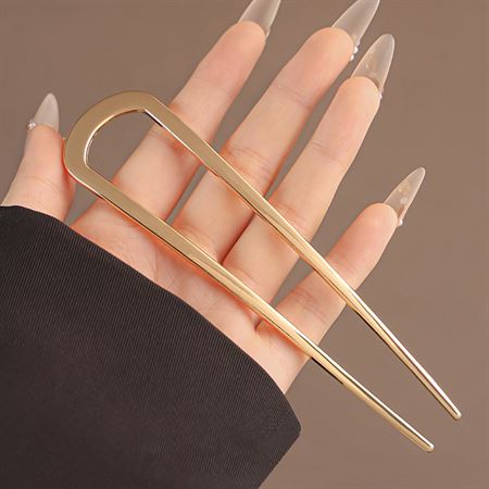 En hånd med lange, spidse negle holder SOHO Atlas XL Hårnål – Guld, en stor guldfarvet hårnål i metal med to ben, frem mod en ensfarvet baggrund – perfekt til at løfte din samling af hårtilbehør fra SOHO.