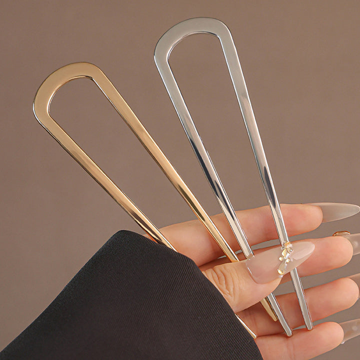 En hånd med lange, pyntede negle holder en guldhårnål og SOHO Atlas XL Hårnål – Sølv – en elegant sølvhårnål til langt hår – mod en ensfarvet brun baggrund.