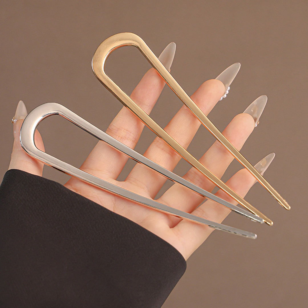 En hånd med lange, mandelformede negle holder to store metalhårnåle – én i guld og én SOHO Atlas XL Hårnål – Sølv – mod en ensfarvet brun baggrund, hvilket fremhæver de elegante håraccessories fra SOHO.