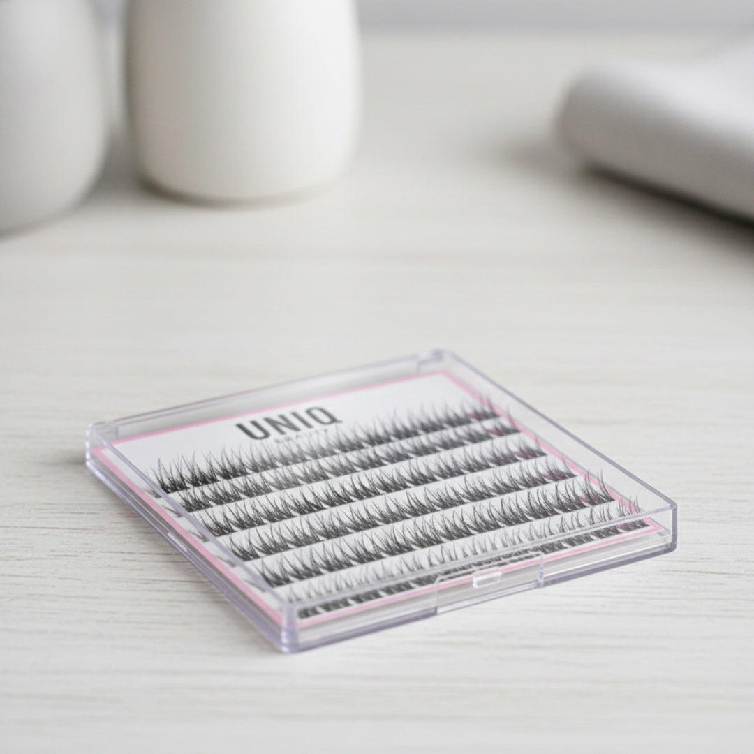 UNIQ Lash Extension Starter Kit - Vippeforlængelse med Cluster Lashes & Bond & Seal fra Stylemekka.dk ligger i sin plastikæske på en lys træoverflade, der er ideel til ethvert DIY-vippeforlængelsessæt, med slørede hvide objekter i baggrunden.