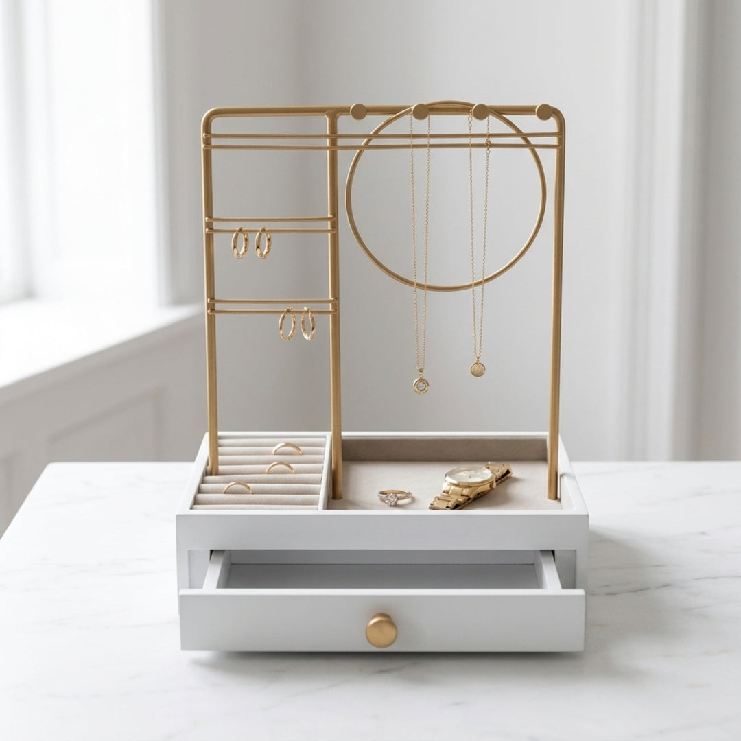 UNIQ Smykkestativ / Smykkeskrin i guld med skuffe – Holder til øreringe, ringe og halskæder (29 cm) fra UNIQ står elegant på en marmoroverflade og fremviser ringe, øreringe, halskæder og et ur, mens skuffen står let på klem. Med blødt oplyste hvide vægge og et vindue i baggrunden tilfører denne praktiske og stilfulde smykkeopbevaring et sofistikeret præg til ethvert rum.