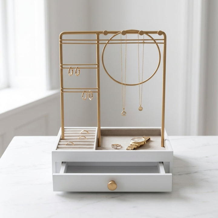 UNIQ Smykkestativ / Smykkeskrin i guld med skuffe – Holder til øreringe, ringe og halskæder (29 cm) fra UNIQ står elegant på en marmoroverflade og fremviser ringe, øreringe, halskæder og et ur, mens skuffen står let på klem. Med blødt oplyste hvide vægge og et vindue i baggrunden tilfører denne praktiske og stilfulde smykkeopbevaring et sofistikeret præg til ethvert rum.
